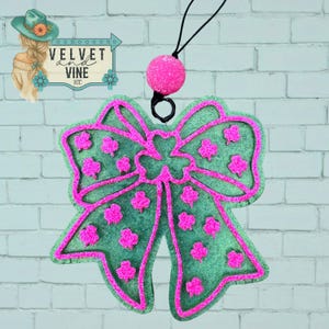 May include: A green bow-shaped air freshener with pink glitter trim and shamrock accents. The air freshener is hanging from a black string and has a pink ball at the top. The image also includes the text "VELVET and VINE ICC".