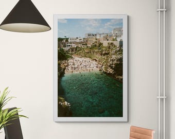 May include: A framed photograph of a coastal scene. The image captures a beach with numerous people, nestled between cliffs and buildings. The water is a clear turquoise, and the sky is blue with clouds. The photograph is displayed in a white frame.