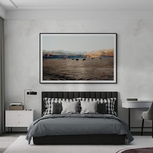 May include: A black framed photograph of a field with a herd of cattle grazing in the foreground. The mountains are in the background. The photograph is hanging on a wall above a gray upholstered bed with black and white checkered pillows.