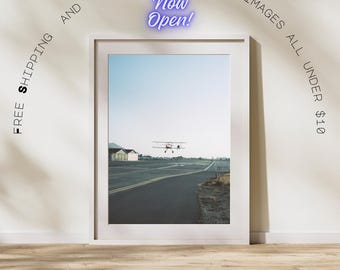 Peut inclure: Photographie encadrée d'un biplan décollant d'une piste d'atterrissage. L'image est prise contre un ciel bleu clair. Les mots "Now Open!" sont dans une police de style néon au-dessus de l'image encadrée.