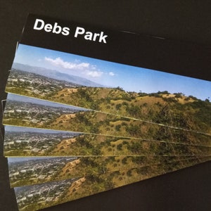 May include: A stack of postcards featuring a scenic view of Debs Park. The cards display a landscape with rolling hills, lush greenery, and a clear blue sky. The text "Debs Park" is printed in white at the top.