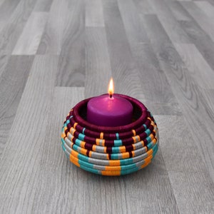 May include: A purple candle with a flame burning inside a woven candle holder with teal, orange, and silver stripes.