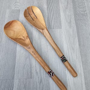 Puede incluir: Un juego de dos cucharas para ensalada de madera con detalles decorativos de cuentas. Las cucharas están hechas de madera natural y tienen un color marrón claro.
