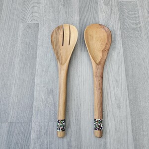 Puede incluir: Un par de utensilios de cocina de madera, una cuchara ranurada y una cuchara maciza, con mangos de color marrón claro y detalles de cuentas en verde, rojo y negro. Las cucharas están hechas de madera clara y se muestran sobre una superficie de madera gris.