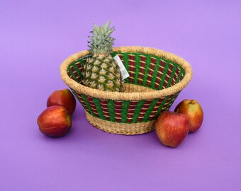 Puede incluir: Una cesta tejida con rayas verdes y rojas contiene una piña y cuatro manzanas rojas. La cesta está sobre una superficie morada.