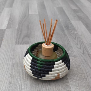 Könnte beinhalten: Ein dekorativer Schilf-Diffusor mit einem gewebten Korb. Der Korb hat ein geometrisches Muster in Schwarz, Weiß, Grün und Beige. Der Diffusor hat einen Holzaufsatz und mehrere braune Schilfrohre. Der Hintergrund ist ein grauer Holzboden.