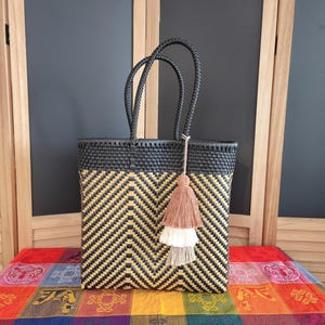 May include: A rectangular woven tote bag featuring a black and tan chevron pattern. The bag has a solid black top border, two braided black handles, and a decorative three-tiered tassel in shades of dusty rose, cream, and light beige hanging from one handle.