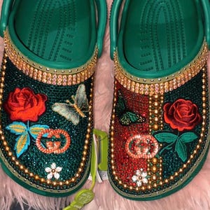 May include: A pair of green clogs embellished with red and green rhinestones, gold trim, and embroidered roses. One clog features a butterfly and the other a butterfly. The clogs have a decorative logo.
