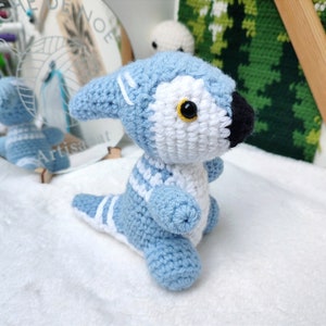 Puede incluir: Un animal de peluche azul y blanco tejido a crochet con una nariz negra y ojos amarillos. El animal está de pie sobre una superficie blanca.