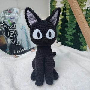 Peut inclure: Peluche de chat noir en crochet avec des yeux blancs et des oreilles grises. Le chat est assis sur une surface blanche.