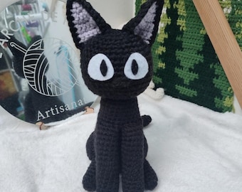 May include: Black crocheted cat plush toy with white eyes and gray ears. The cat is sitting on a white surface.