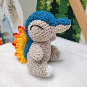 Può includere: Peluche all'uncinetto di un personaggio Pokemon blu e grigio con una coda a forma di fiamma gialla e arancione.