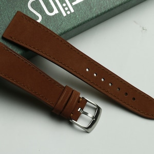 May include: A brown leather watch strap with stitching and a silver buckle.