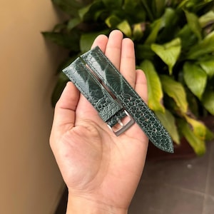 May include: A dark green watch strap featuring a textured, scale-like pattern and a polished silver-tone metal buckle. The strap is composed of two pieces, with visible stitching along the edges and multiple adjustment holes for a secure fit.