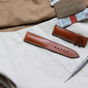 May include: Two brown leather watch straps with white stitching. One strap is longer than the other and has a buckle. A silver watch repair tool is also in the image.