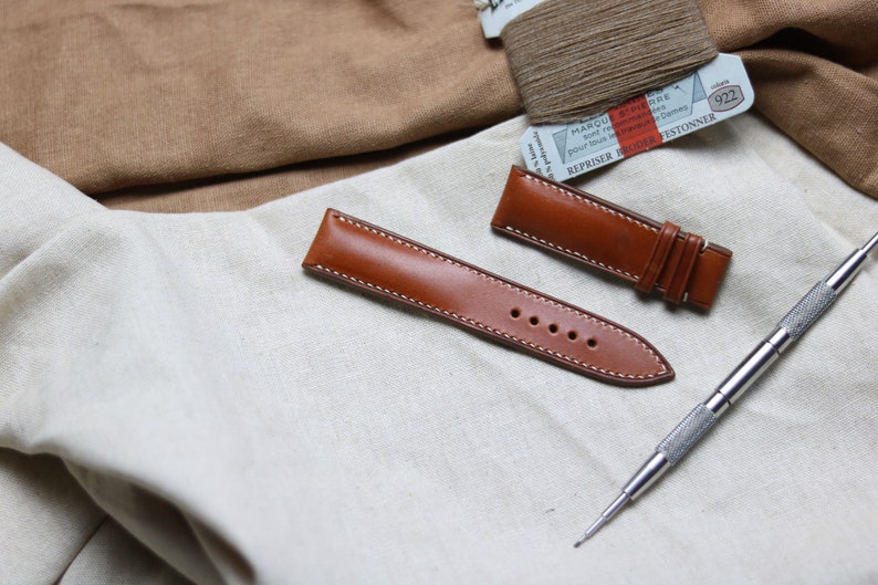 May include: Two brown leather watch straps with white stitching. One strap is longer than the other and has a buckle. A silver watch repair tool is also in the image.