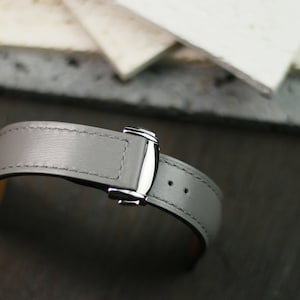 Puede incluir: Una correa de reloj de cuero gris con una hebilla plateada.