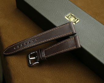 May include: Two brown leather watch straps with stitching. The straps are on a brown surface and a green box with the word "SUPA" on it.
