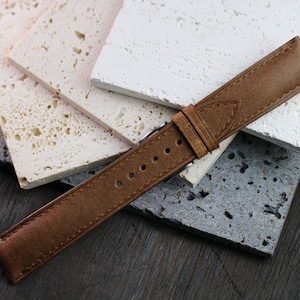May include: A brown leather watch strap with a silver buckle. The strap is shown against a backdrop of textured, light-colored stone tiles. The watch band has several holes for adjustment.