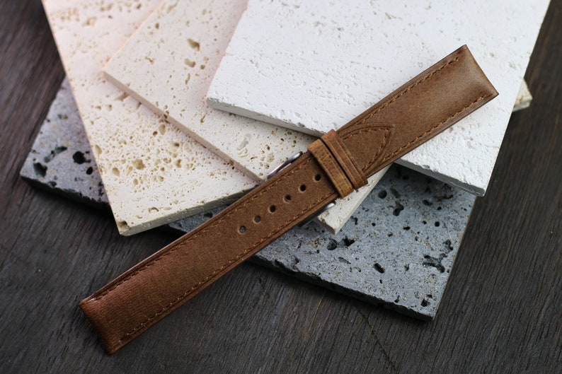 May include: A brown leather watch strap with a silver buckle. The strap is shown against a backdrop of textured, light-colored stone tiles. The watch band has several holes for adjustment.