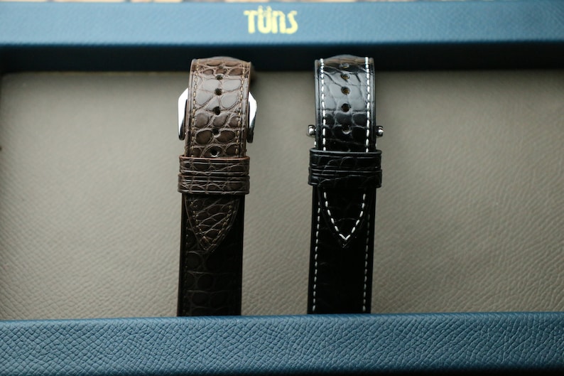 Peut inclure: Deux bracelets de montre, l'un brun et l'autre noir, sont pr&eacute;sent&eacute;s sur un fond bleu. Les bracelets sont en cuir et pr&eacute;sentent un design cousu.