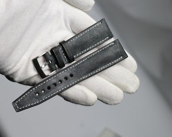 May include: A black leather watch strap with white stitching. The strap has a buckle and is shown on a white surface.