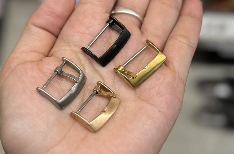 K&ouml;nnte beinhalten: Vier Uhrenarmband-Schlie&szlig;en in verschiedenen Ausf&uuml;hrungen: Silber, Schwarz, Gold und Ros&eacute;gold. Die Schlie&szlig;en sind alle gleich gro&szlig; und haben die gleiche Form, mit einem einfachen, rechteckigen Design.