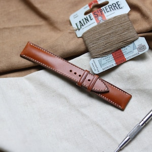 May include: A brown leather watch strap with white stitching. The strap is lying on a white fabric surface. There is a spool of brown thread and a tool for watch strap repair in the background.