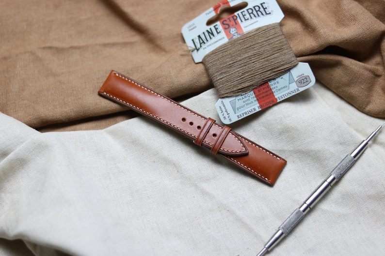 May include: A brown leather watch strap with white stitching. The strap is lying on a white fabric surface. There is a spool of brown thread and a tool for watch strap repair in the background.