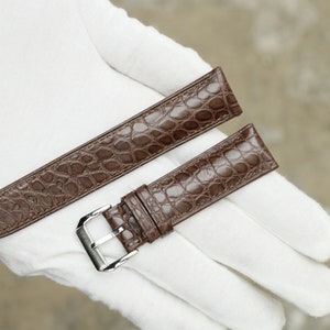 Peut inclure: Deux bracelets de montre en cuir marron avec un motif crocodile. Le bracelet de gauche est plus long et a une extr&eacute;mit&eacute; pointue. Le bracelet de droite est plus court et a une boucle.