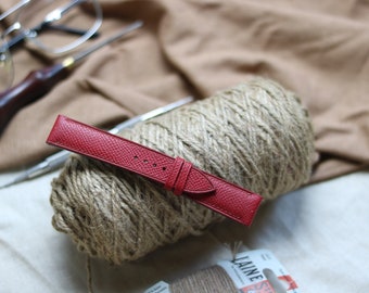 May include: A red leather watch strap with a smooth texture. The strap is lying on top of a ball of twine.