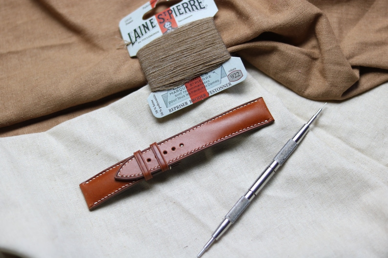 May include: A brown leather watch strap with white stitching, a small spool of brown thread, and a silver watch tool on a white fabric background.