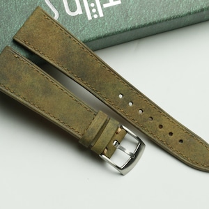 Pode incluir: Uma pulseira de relógio de couro verde com uma fivela prateada. A pulseira é feita de couro envelhecido e tem uma borda costurada.