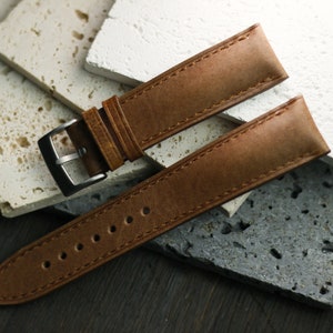 May include: A brown leather watch strap with a silver buckle. The strap is made of textured leather and has visible stitching. The watch band is displayed on a surface of stone tiles.