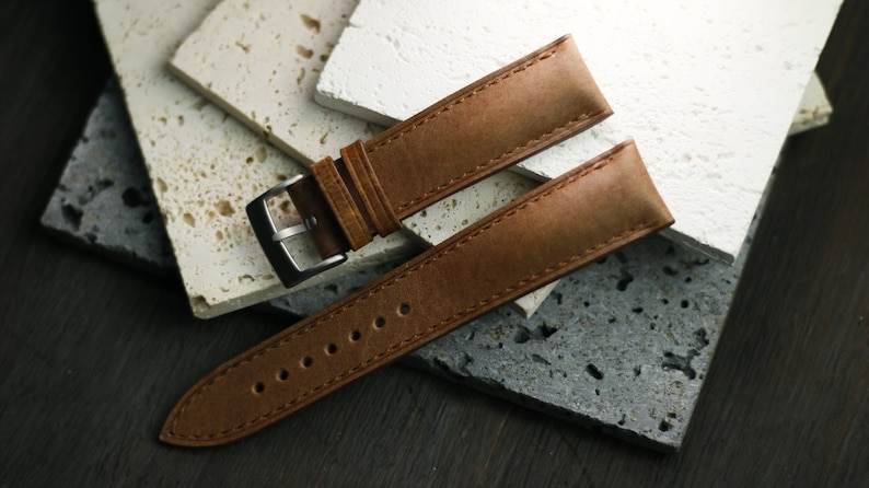 May include: A brown leather watch strap with a silver buckle. The strap is made of textured leather and has visible stitching. The watch band is displayed on a surface of stone tiles.