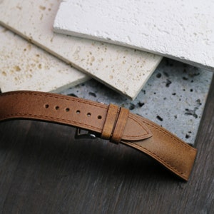 May include: A brown leather watch strap with a stainless steel buckle. The strap has several holes for adjustment and is set against a backdrop of textured stone tiles and a dark wood surface.