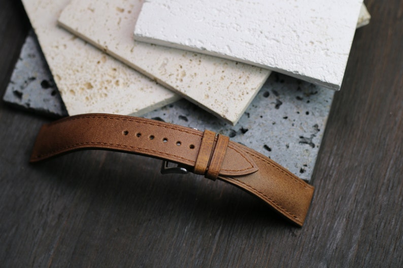 May include: A brown leather watch strap with a stainless steel buckle. The strap has several holes for adjustment and is set against a backdrop of textured stone tiles and a dark wood surface.