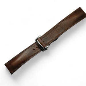 May include: A brown leather watch strap with a silver-colored buckle. The strap is made of textured leather and has a rectangular shape. The watch band is angled across a white background.