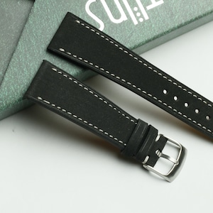 Può includere: Due cinturini per orologio in pelle nera con cuciture bianche e fibbie argentate. I cinturini sono disposti su una superficie bianca.