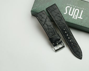 May include: Two black leather watch straps with white stitching. The straps are on a white surface and a green box with the text "Tuns Leather Strap" is in the background.
