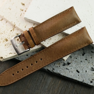 May include: A brown leather watch strap with a silver buckle. The strap is made of textured leather and has several holes for adjustment. The watch strap is displayed on a textured surface.