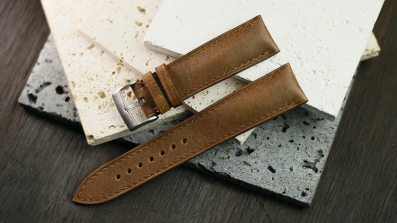 May include: A brown leather watch strap with a silver buckle. The strap is made of textured leather and has several holes for adjustment. The watch strap is displayed on a textured surface.