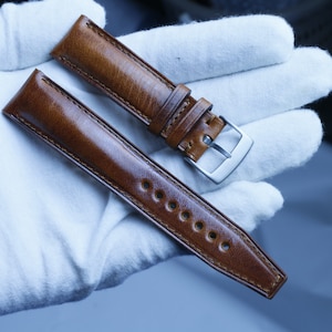 Puede incluir: Dos correas de reloj de cuero marrón con detalles de costura. Una correa tiene una hebilla plateada.