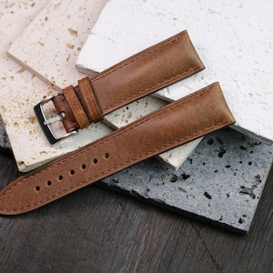 May include: A brown leather watch strap with a silver buckle, lying on a textured surface. The strap has visible stitching and several holes for adjustment. The background features various stone tiles.