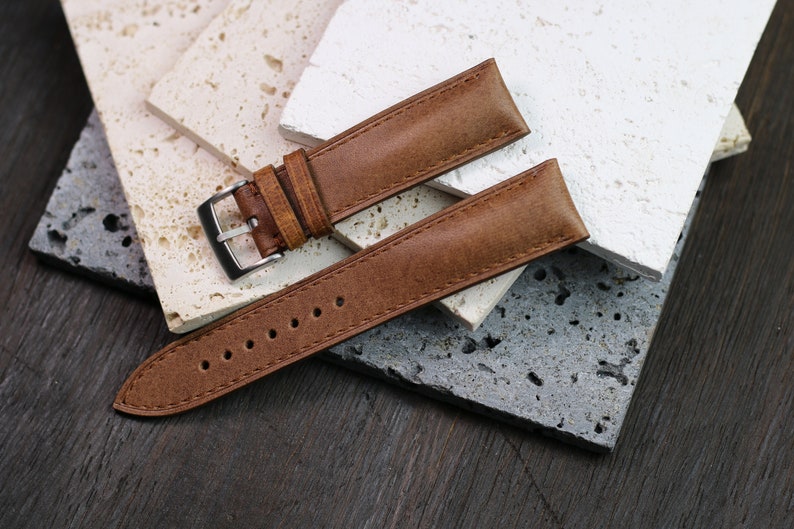 May include: A brown leather watch strap with a silver buckle, lying on a textured surface. The strap has visible stitching and several holes for adjustment. The background features various stone tiles.