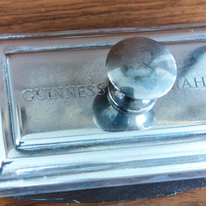 Puede incluir: Un pisapapeles plateado con un asa redondeada. La base rectangular presenta el texto grabado "GUINNESS MAHON". El pisapapeles est&aacute; hecho de un metal brillante y reflectante.