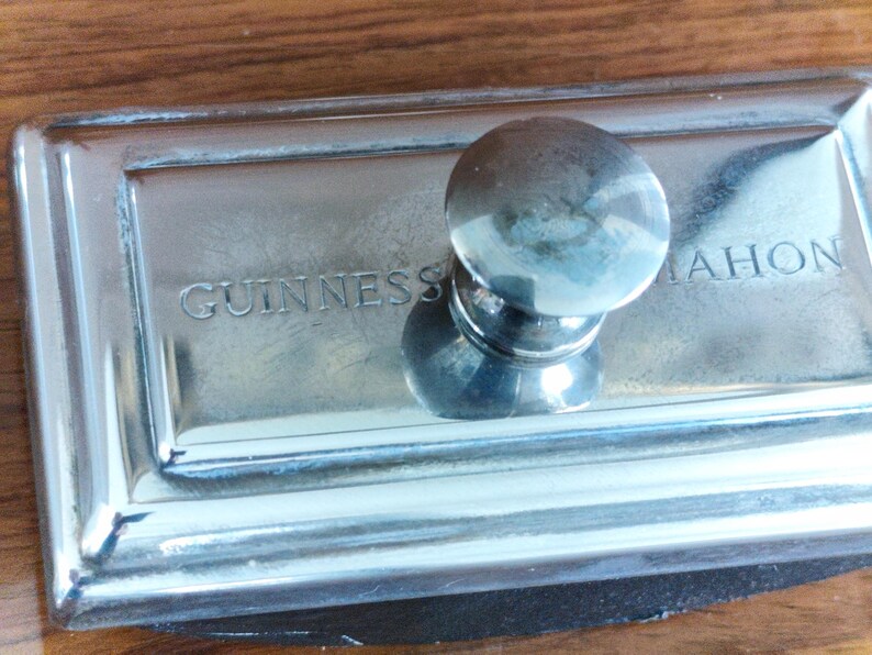 Puede incluir: Un pisapapeles plateado con un asa redondeada. La base rectangular presenta el texto grabado "GUINNESS MAHON". El pisapapeles est&aacute; hecho de un metal brillante y reflectante.
