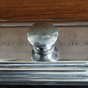 Puede incluir: Un secante de escritorio plateado con un asa redondeada. Las palabras "GUINNESS" y "MAHON" est&aacute;n grabadas en la superficie rectangular. El secante est&aacute; sobre un fondo de madera.