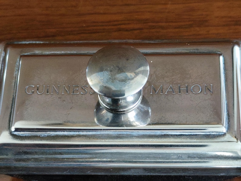 Puede incluir: Un secante de escritorio plateado con un asa redondeada. Las palabras "GUINNESS" y "MAHON" est&aacute;n grabadas en la superficie rectangular. El secante est&aacute; sobre un fondo de madera.