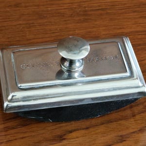 Puede incluir: Un secante de tinta plateado con las palabras "GUINNESS MAHON" grabadas. Tiene un asa redondeada y una base rectangular con una almohadilla negra. El art&iacute;culo est&aacute; sobre una superficie de madera. Mide unos 10 cm de largo.